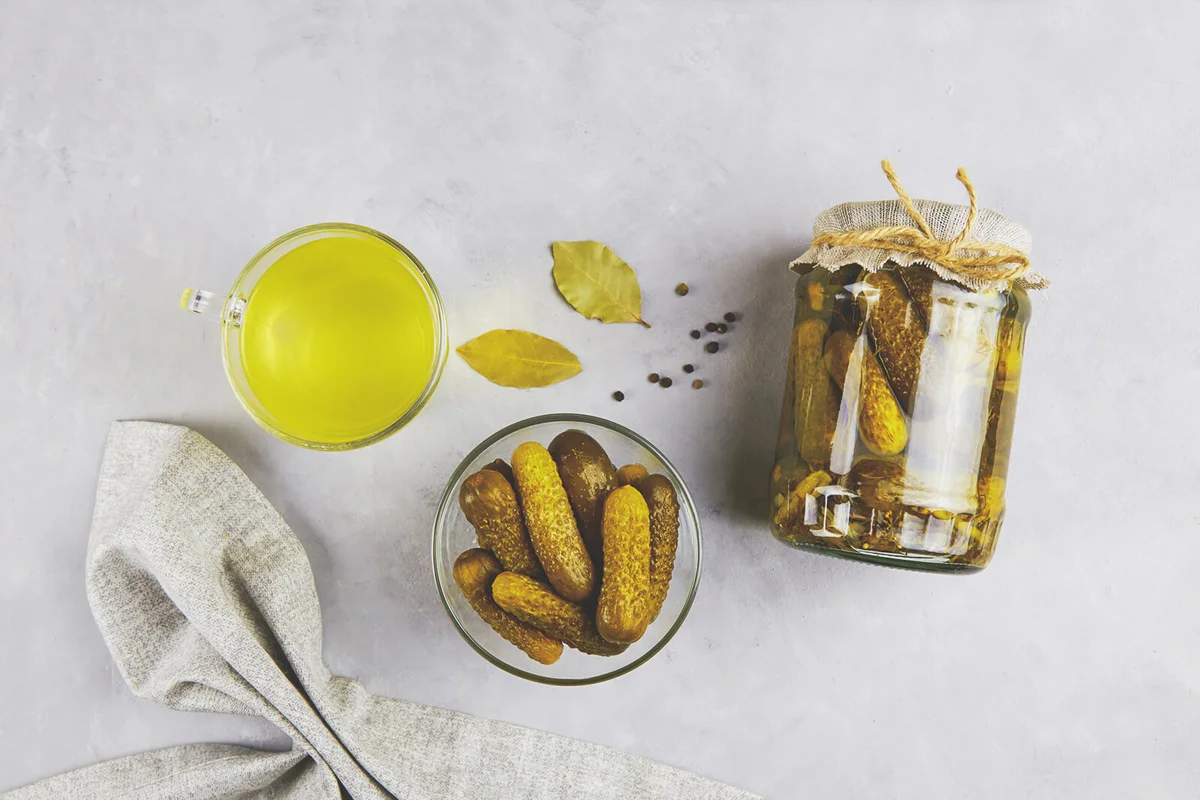 Pickled cucumbers in a glass jar and bowl, pickle juice in glass on a light background