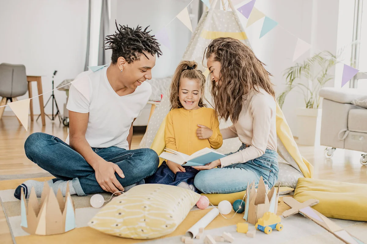 parents reading a book in front of the tent on the floor in the living room to their child