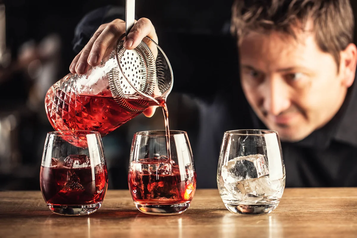 Skilled bartender pours negroni short drink cocktail into three glasses