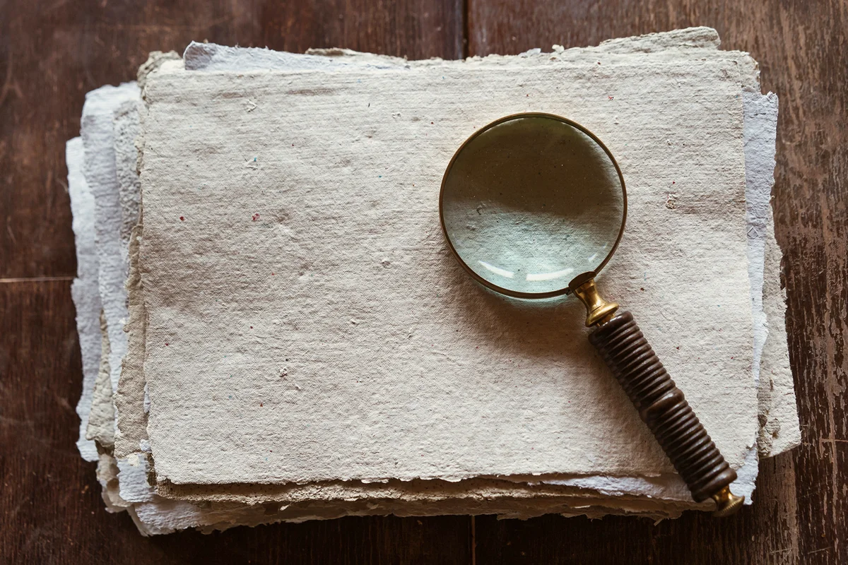 A stack of sheets of recycled paper lying on a wooden table and an antique hand held magnifier glass