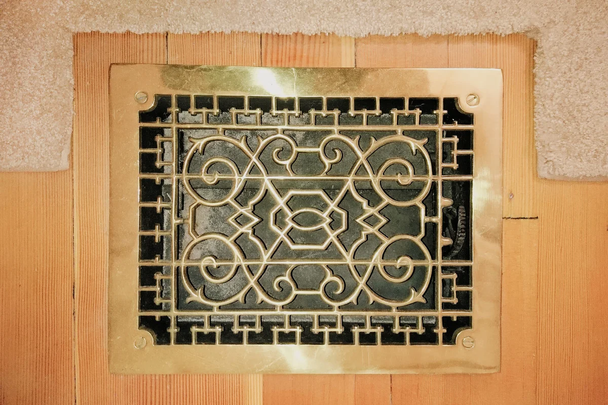 Overhead close-up of an ornate brass air duct vent surrounded by hard wood flooring and a rug