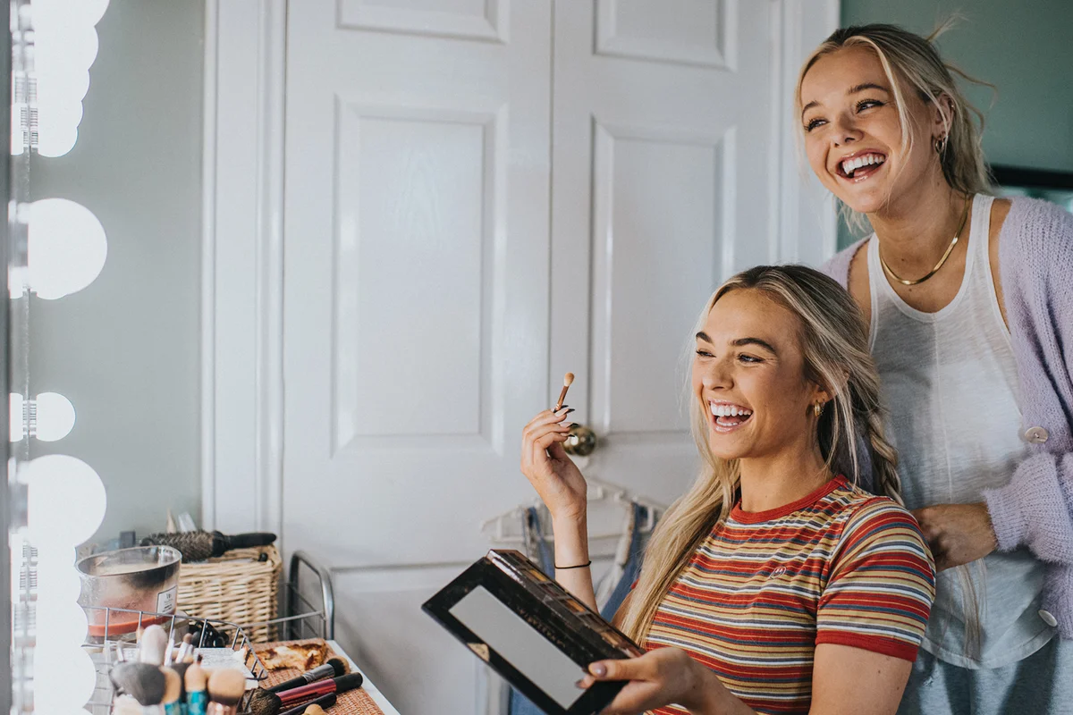 Two laughing young woman front of an illuminated mirror and apply make-up