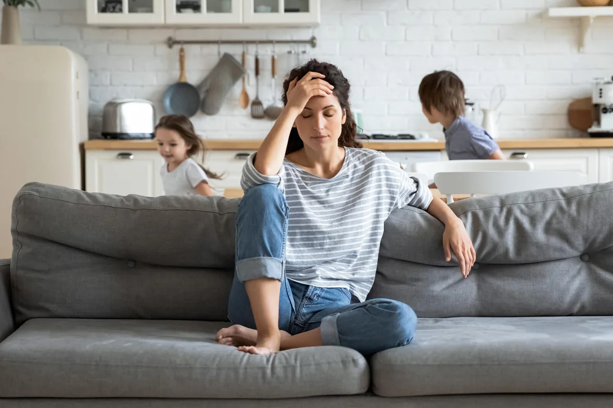 Exhausted mother sitting on couch while kids running at home