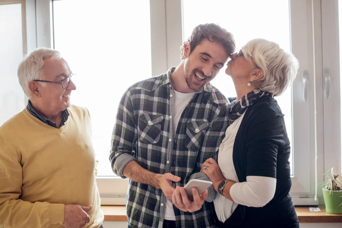 Young man looking at phone, his mother kissing him and father looking at them