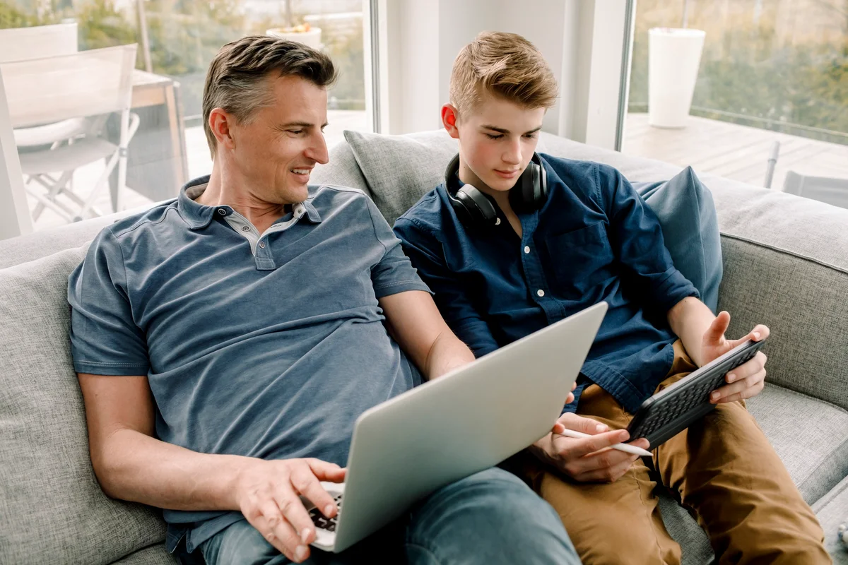 Smiling father with laptop looking at son using digital tablet while sitting on sofa at home