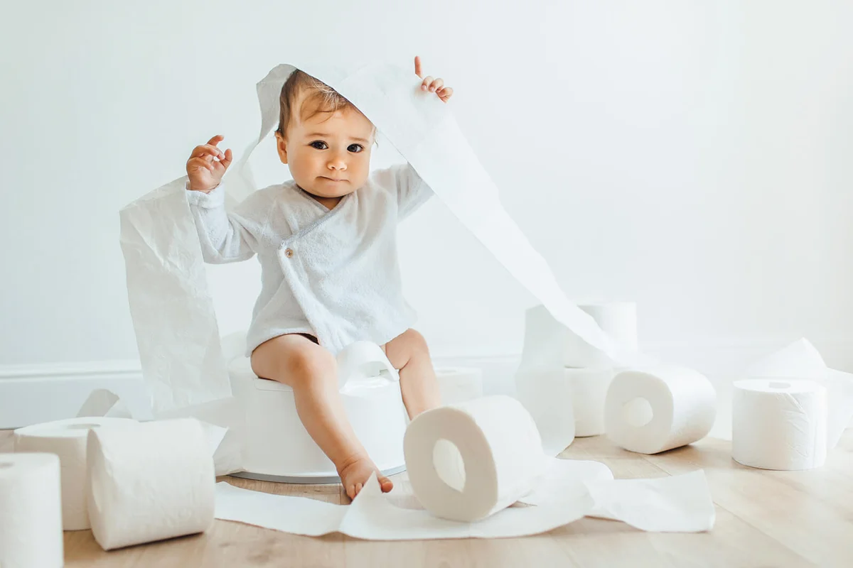 Cute baby girl sitting on white chamber pot with toilet paper rolls
