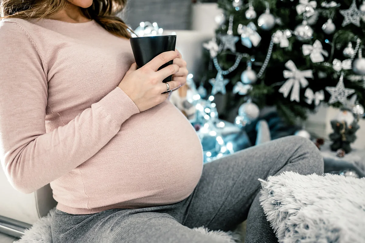 Happy pregnant woman drinking coffee next to the Christmas tree