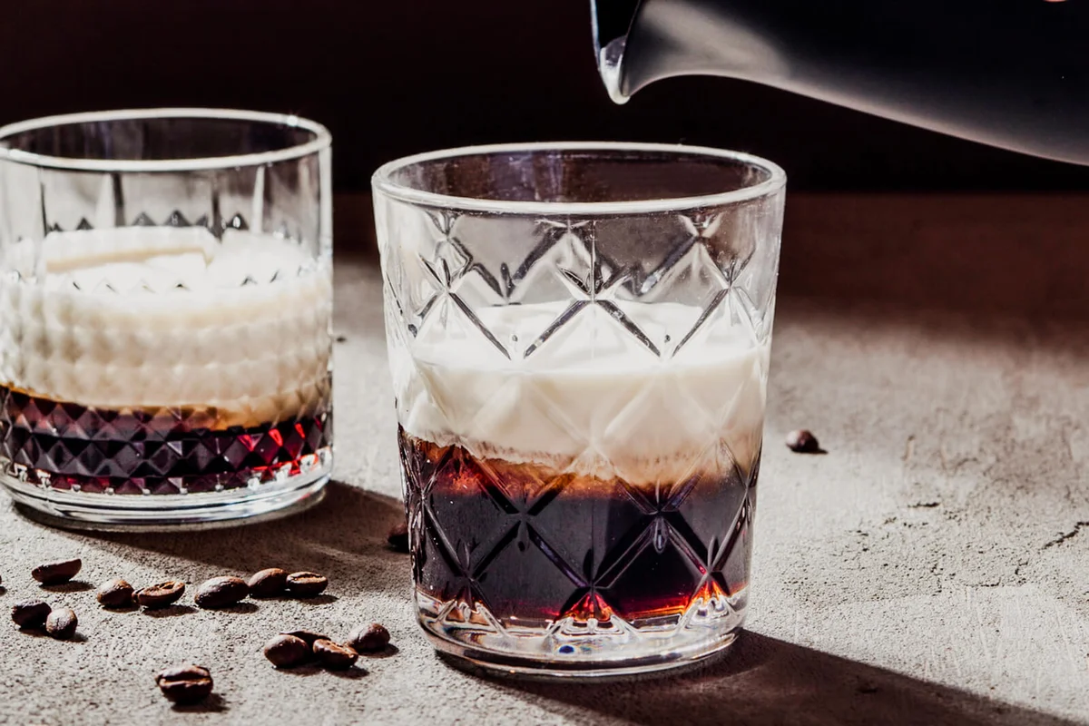 Cropped hand of woman holding pitcher over White Russian cocktail on concrete table