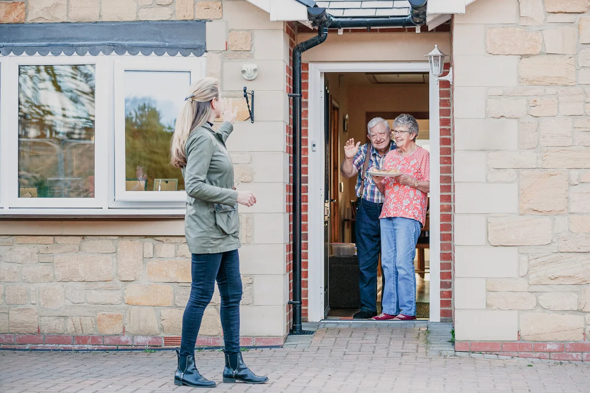 female stands back waving from the front door of a bungalow after delivering food