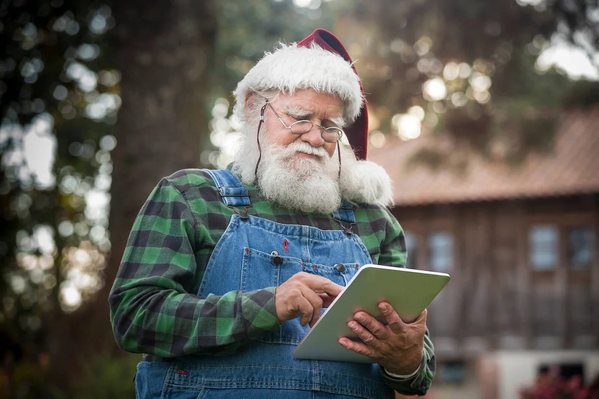 Santa Claus checking his digital tablet in front of his log cabin in the woods