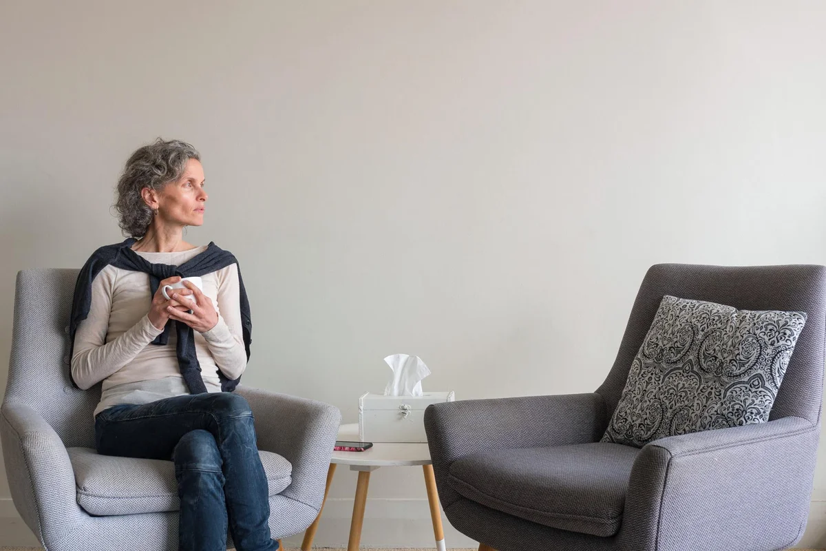 Middle aged woman seated looking pensive in living room with second empty armchair