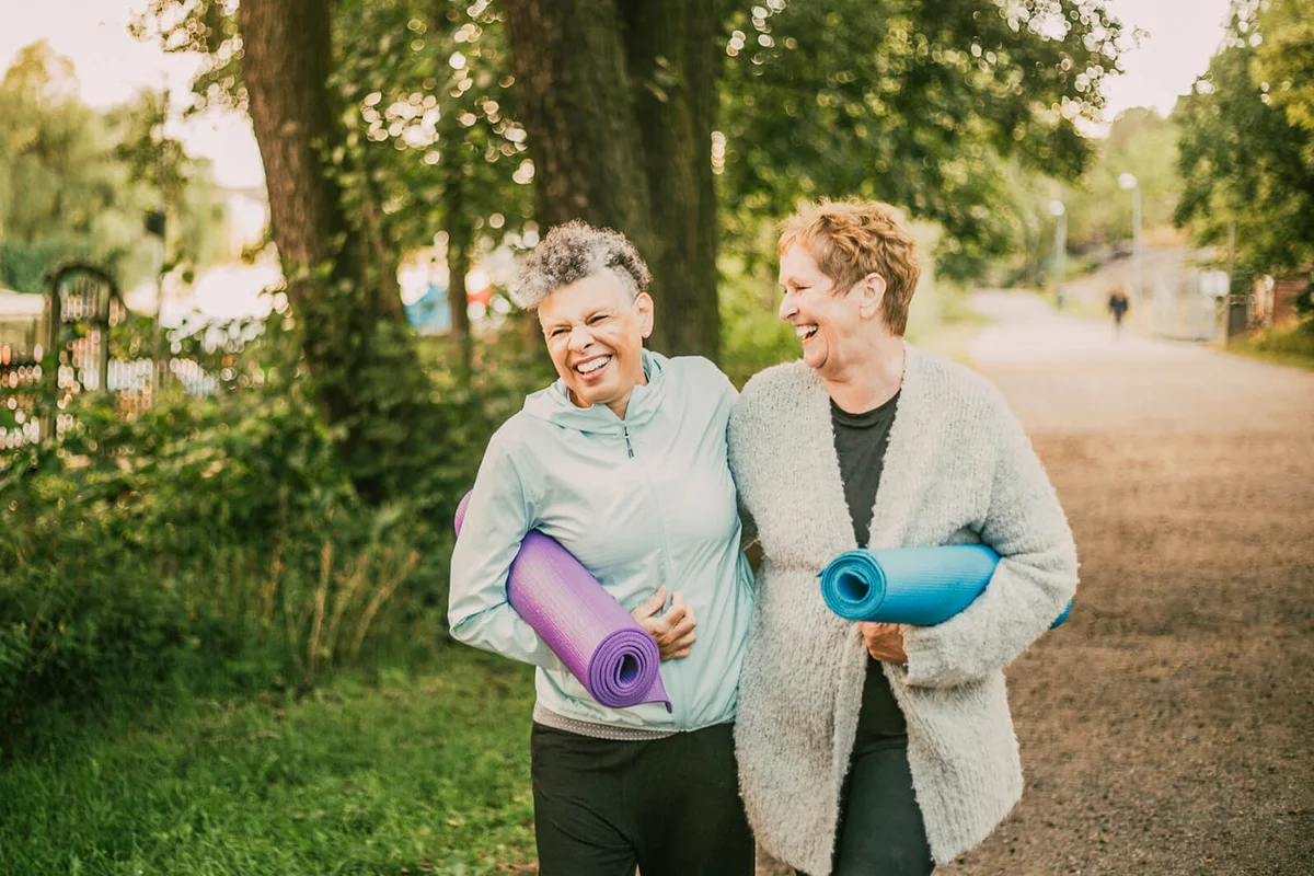Smiling female friends holding exercise mat while walking on footpath