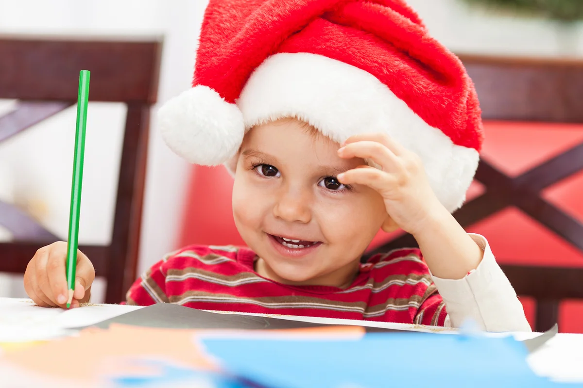 Cute little boy coloring a Christmas card wearing Santa's hat and smiling