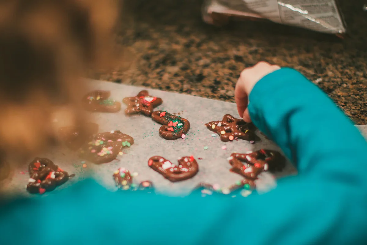 Little girl stands in a kitchen and puts red, green and white sprinkles on chocolate covered pretzels