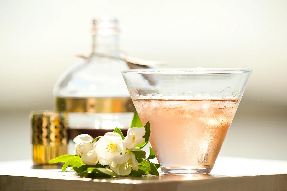 Blueberry martini outside on a table with white flowers and a bottle in background