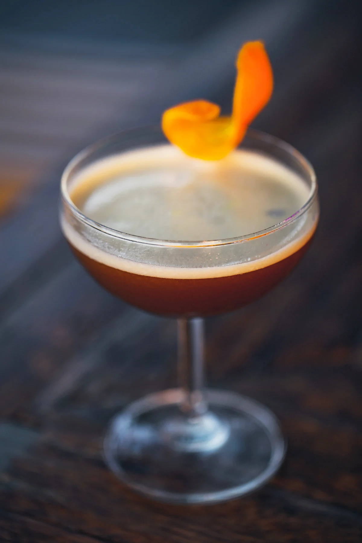 Dark cocktail garnished with an orange twist on a dark bar setting
