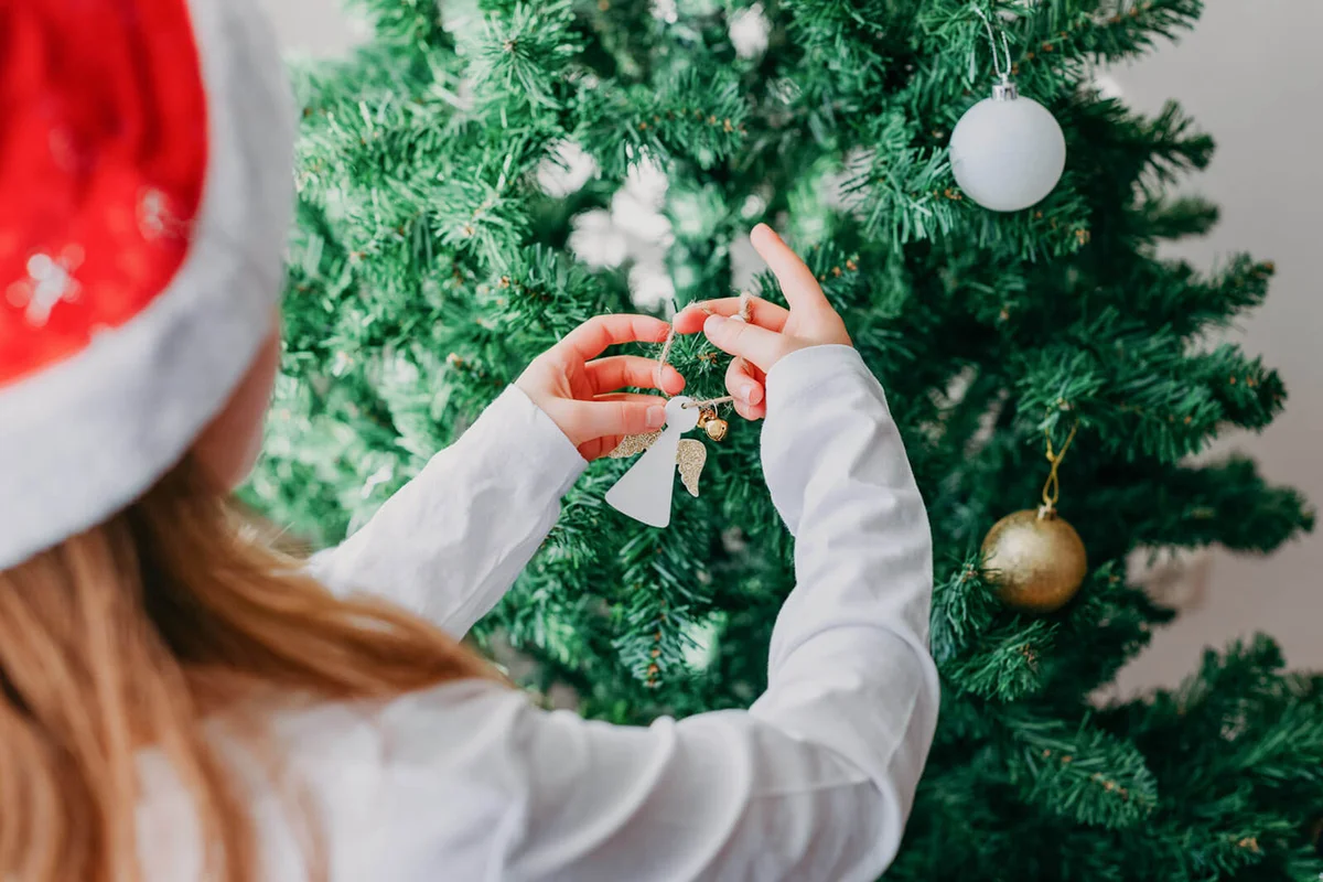 Girl Decorates An Artificial Christmas Tree In A Santa Hat With Christmas Deco
