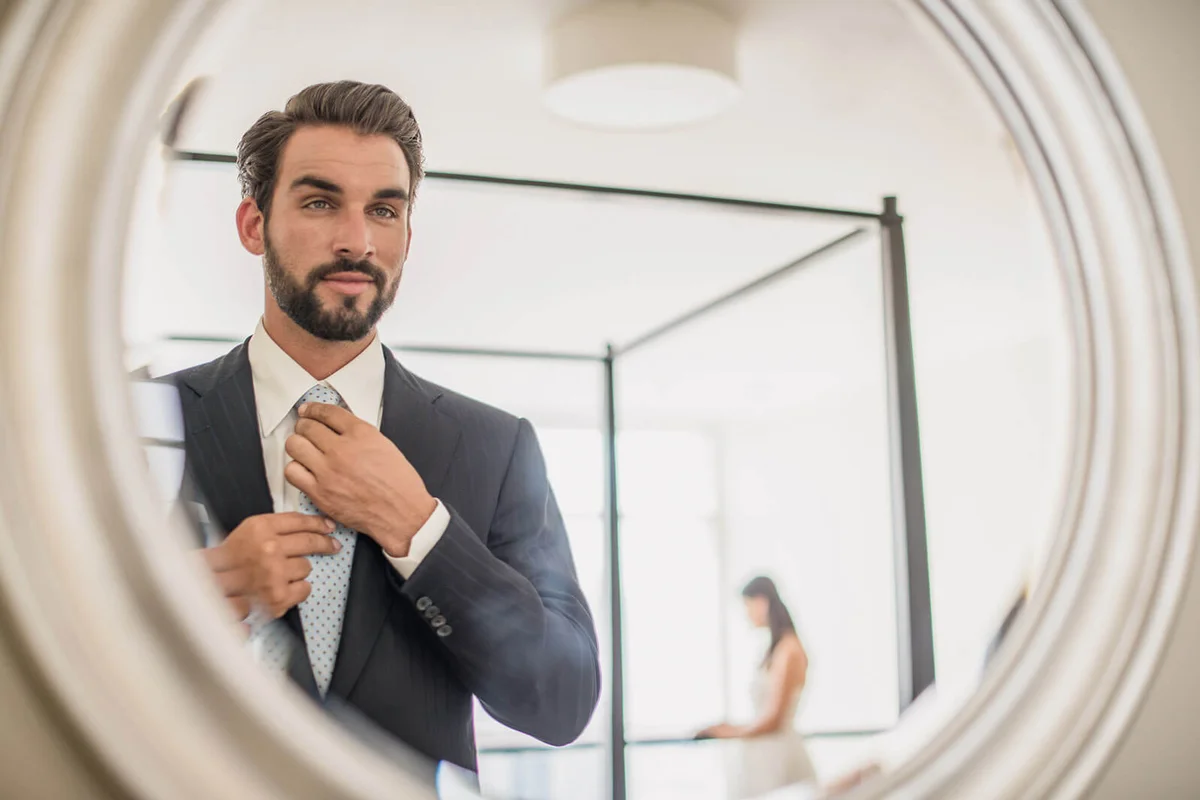 Mirror reflection of young businessman adjusting shirt and tie in hotel room