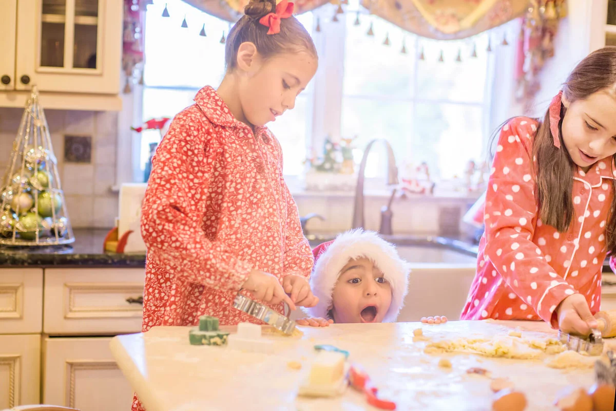 Boy in kitchen wearing santa hat making cookies with sisters peeking over kitchen counter open mouthed