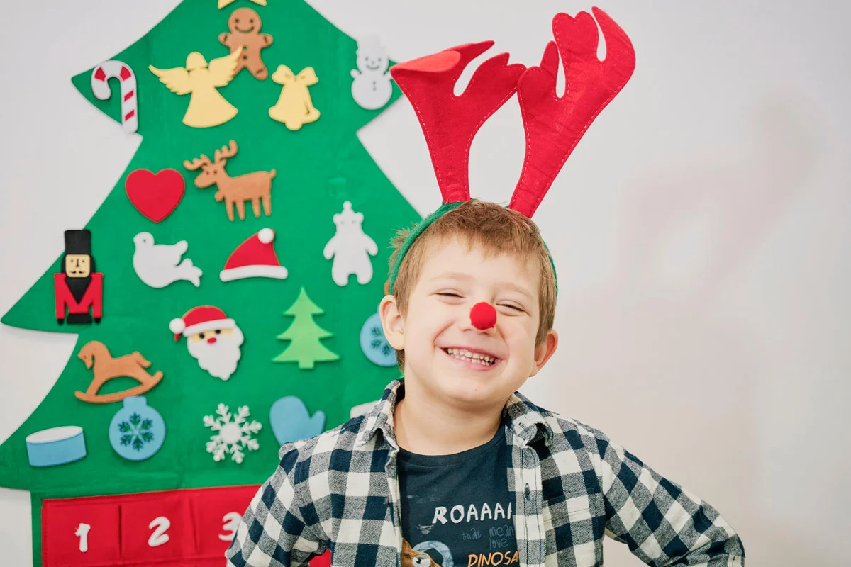 Smiling child posing for a Christmas postcard with reindeer ears and a wall-mounted Christmas tree behind him