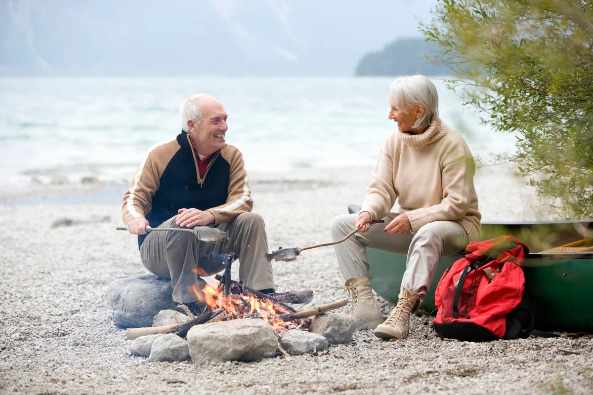 Senior couple sitting at campfire, grilling fish