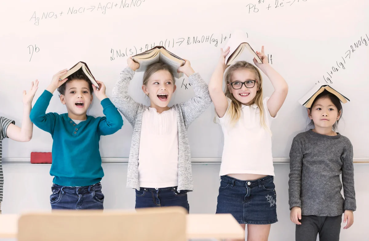 Happy pupils with books above their heads standing at whiteboard with formulas in class