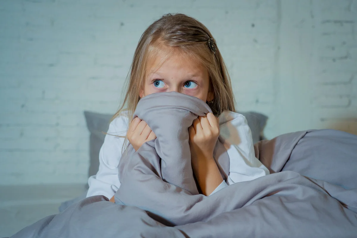 Scared little girl staying sleepless hiding behind the duvet looking horrified