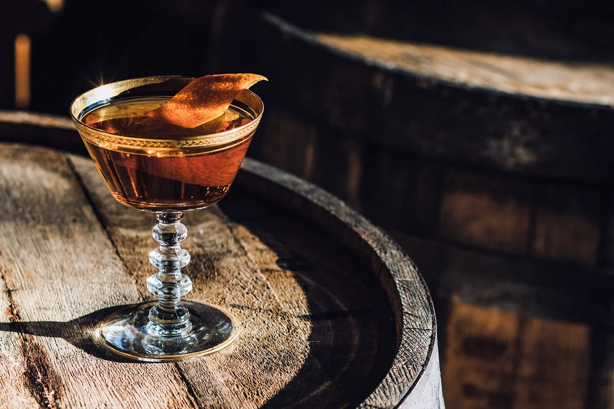 Manhattan Bourbon Whiskey Cocktail In Vintage Crystal Hotel Glass Orange Garnish Atop Wood Barrel