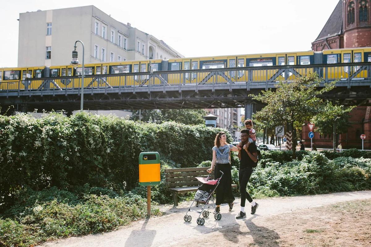 oung man carrying daughter on shoulders while woman pushing baby stroller at footpath against railway bridge