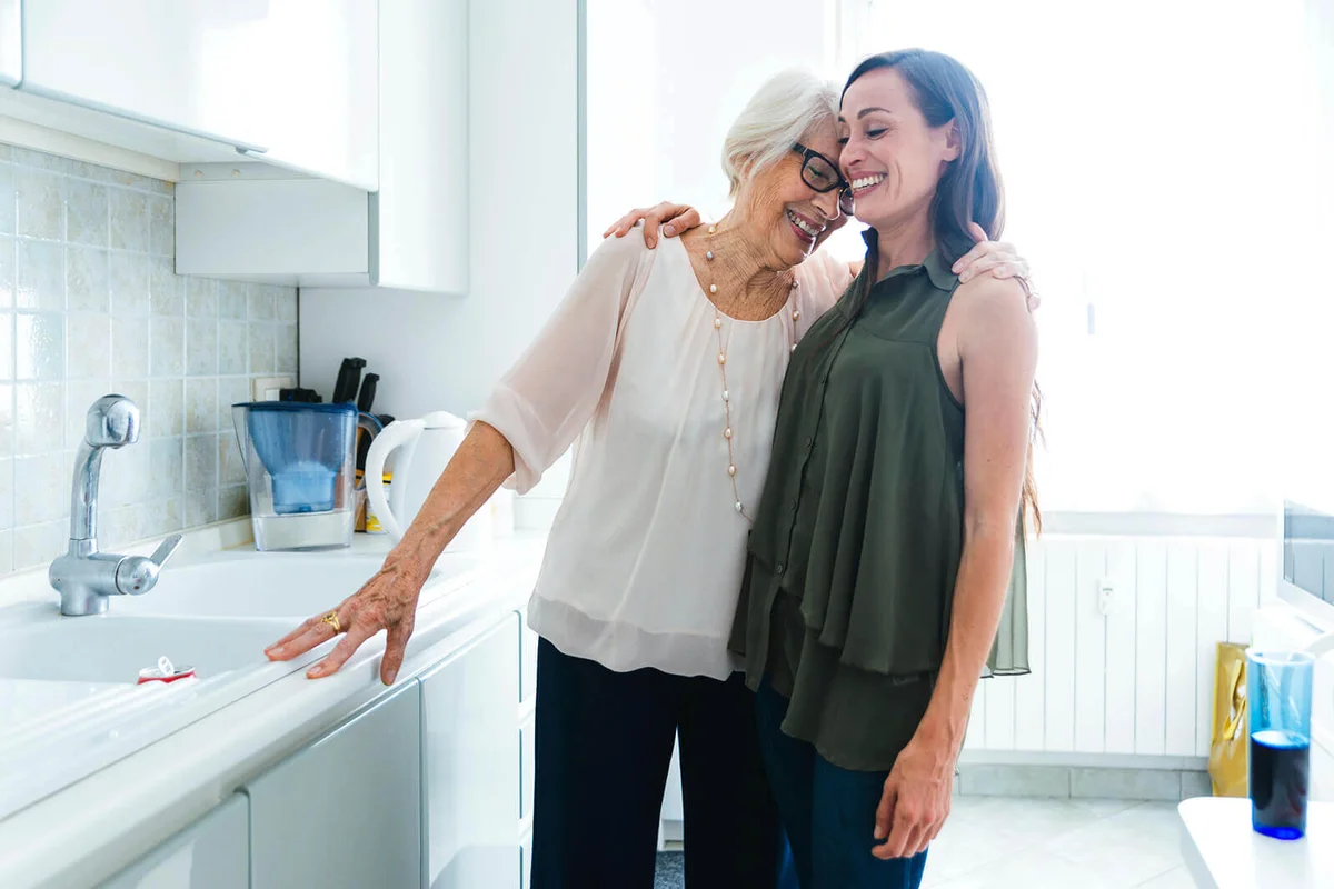 Smiling granddaughter and grandmother standing with arm around while hugging in kitchen at home