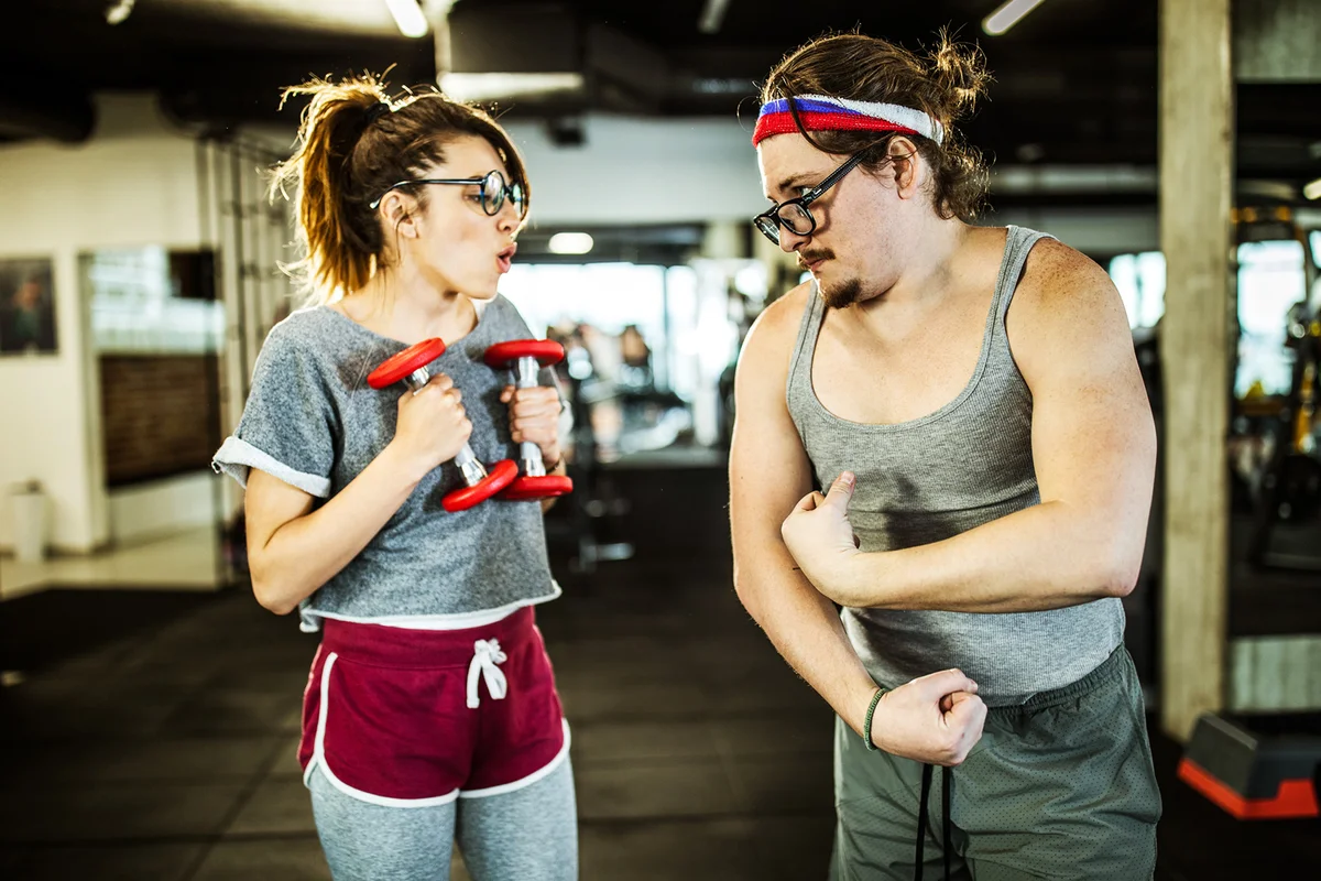 Nerdy male athlete showing off with his biceps in front of his girlfriend in a gym