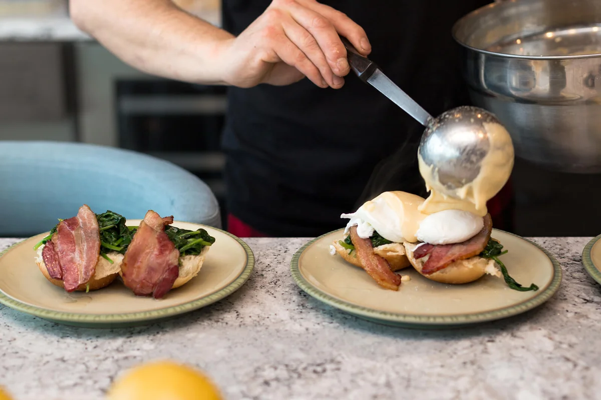 Eggs benedict with crisp bacon and spinach being topped with hollandaise sauce