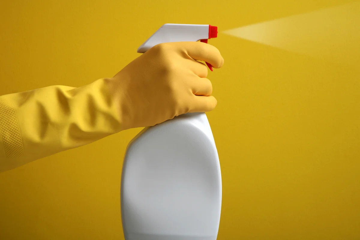 Hand in yellow rubber glove helds a plastic spray bottle with cleaning detergent on a yellow background.