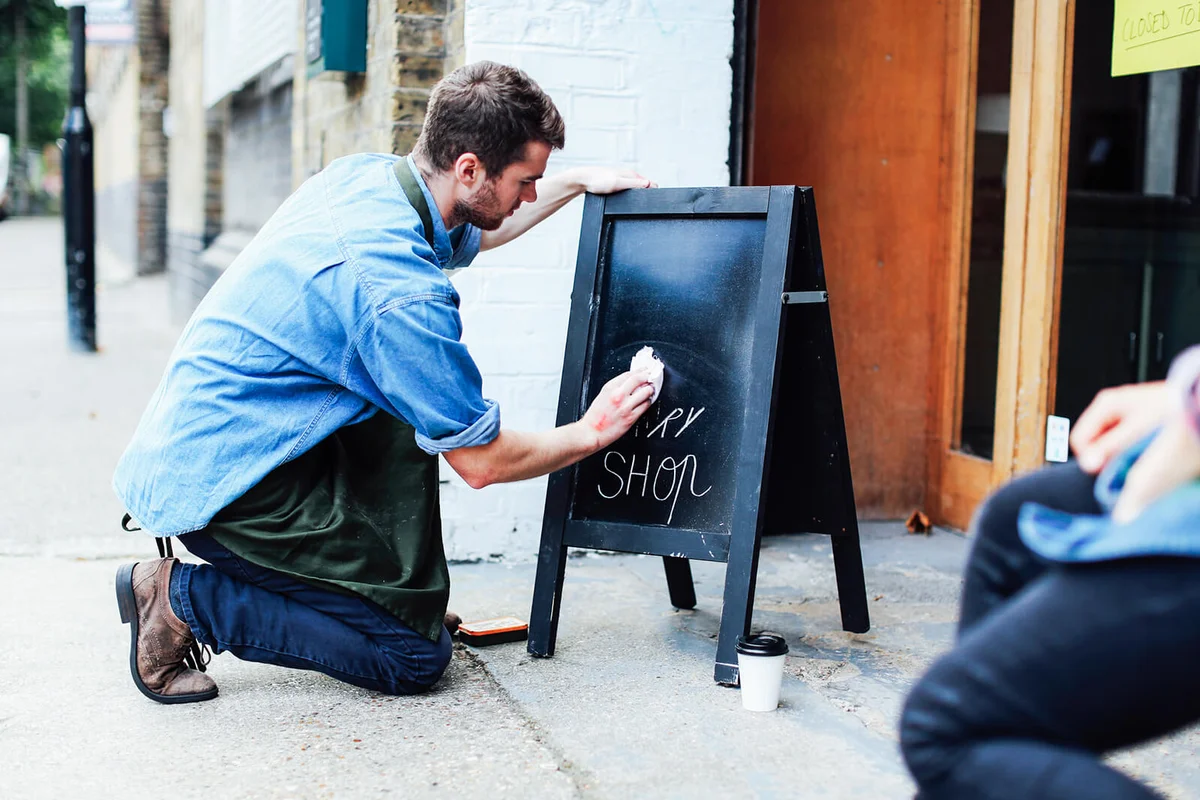 Young man kneeling on pavement and wiping blackboard outside workshop