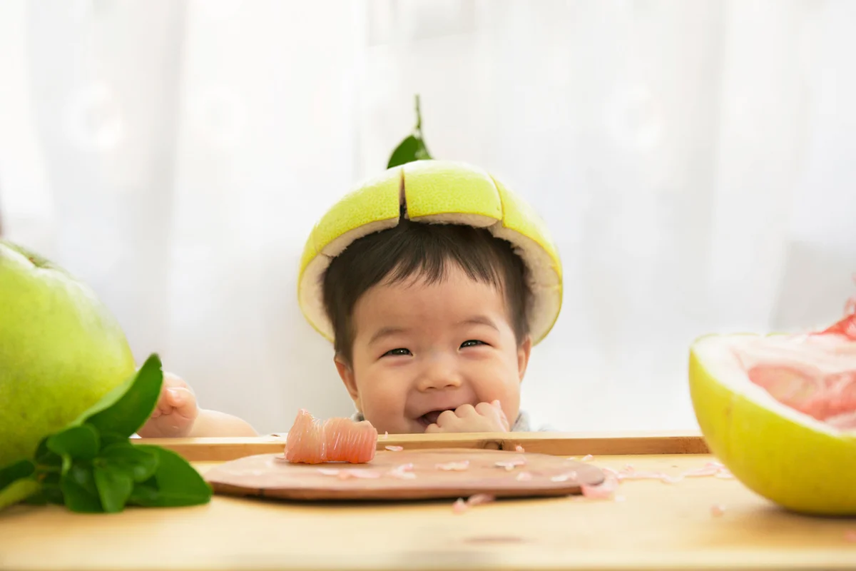 Toddler eating fruits, looking silly with pomelo skin as a hat