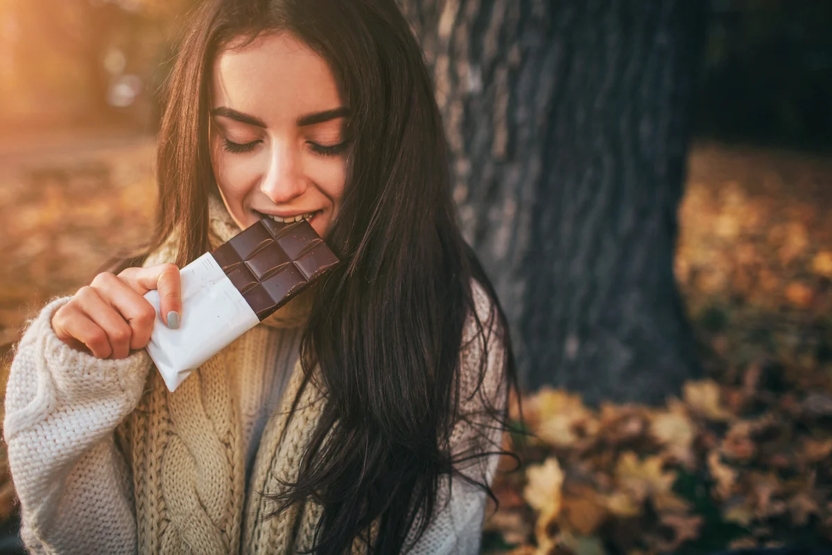 Beautiful young brunette sitting on a autumn day in a park, eating a chocolate bar