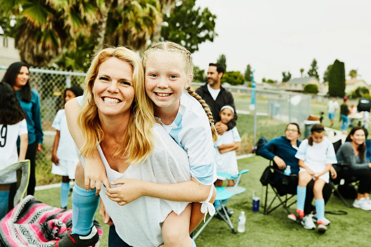 Smiling mother giving daughter piggy back ride on sidelines after soccer game
