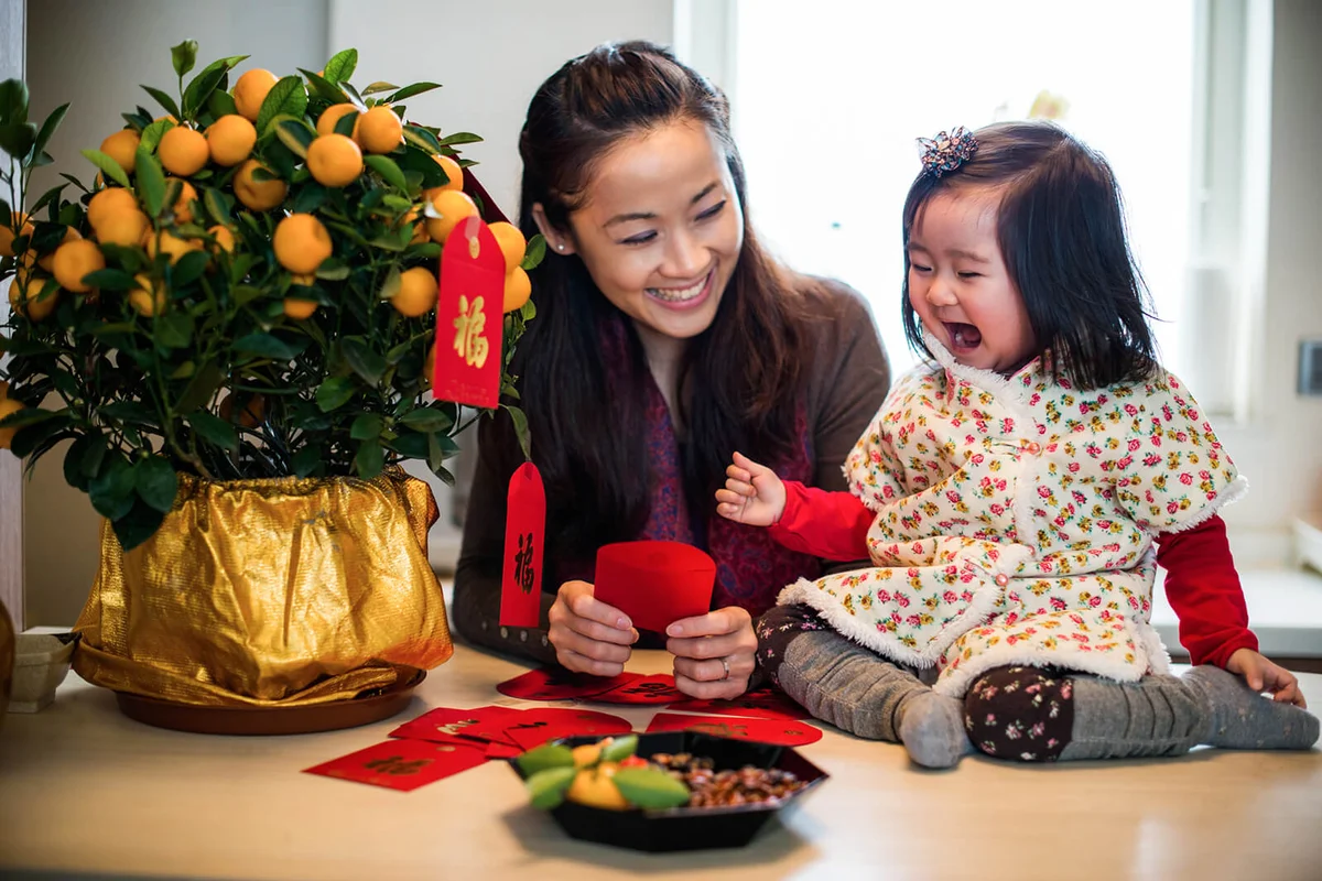 Pretty young mom holding a red envelope decorating traditional tangerine tree for Chinese New Year with toddler girl