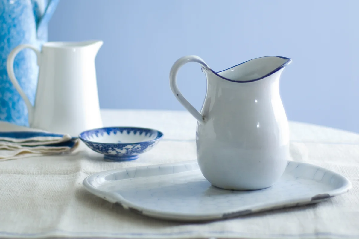 Enameled white pitcher in foreground on enamel platter, with porcelain and enameled pitchers in background.