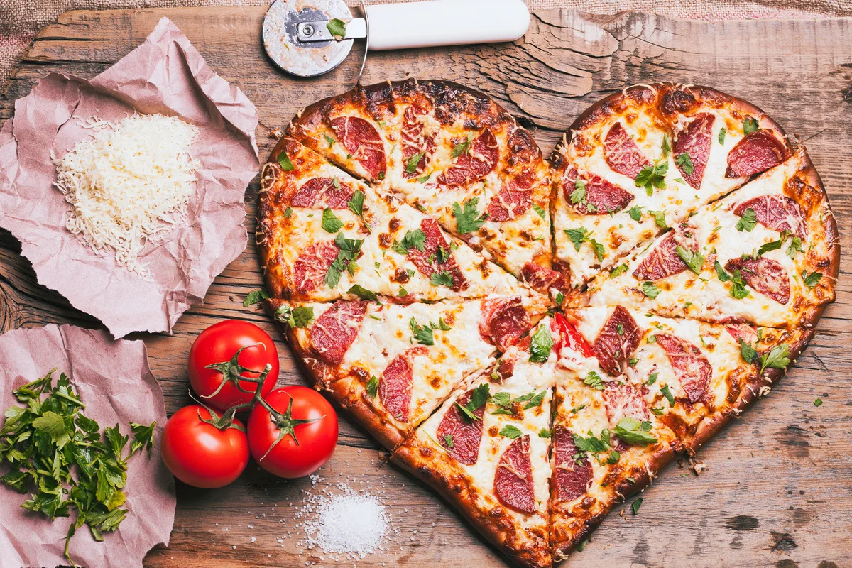Heart-shaped pizza and ingredients on cutting board