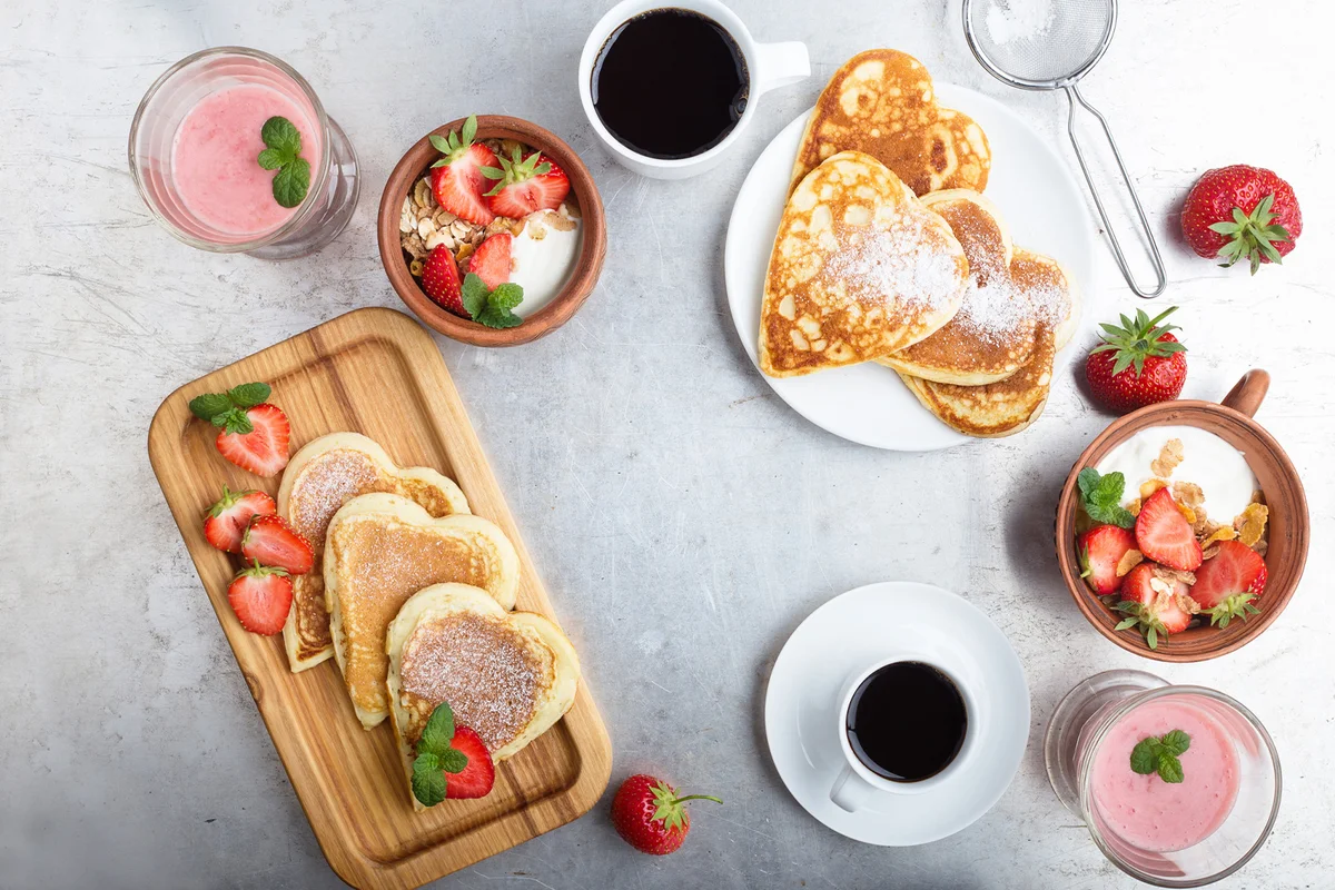 Breakfast table with heart shaped pancakes, strawberries, muesli, fruit smoothies and coffee