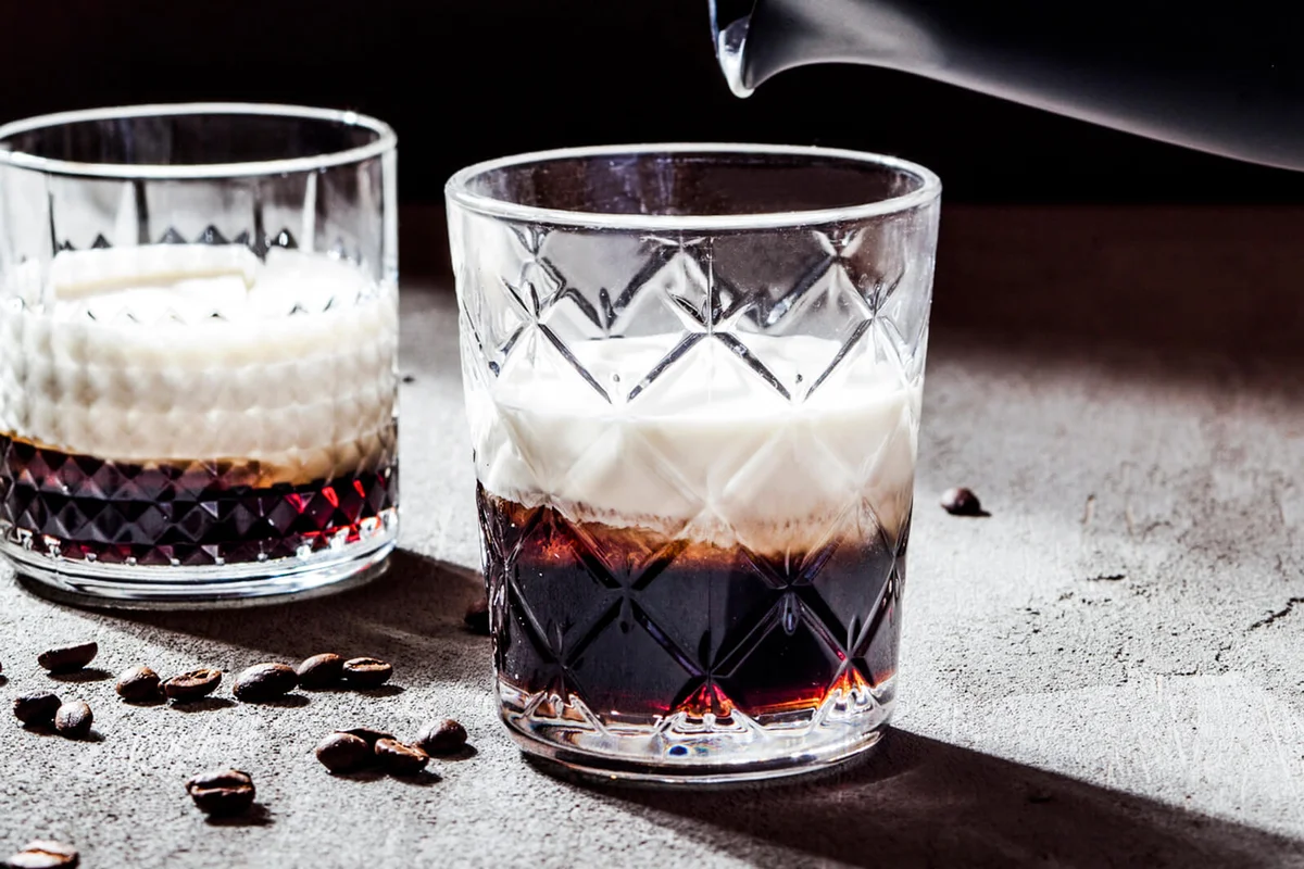Cropped hand of woman holding pitcher over White Russian cocktail on concrete table