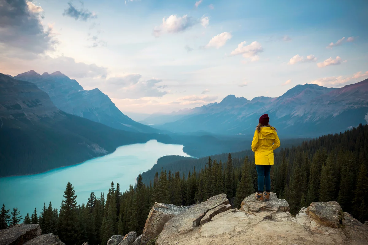 Young woman in a yellow jacket standing on a rock overlooking a lake
