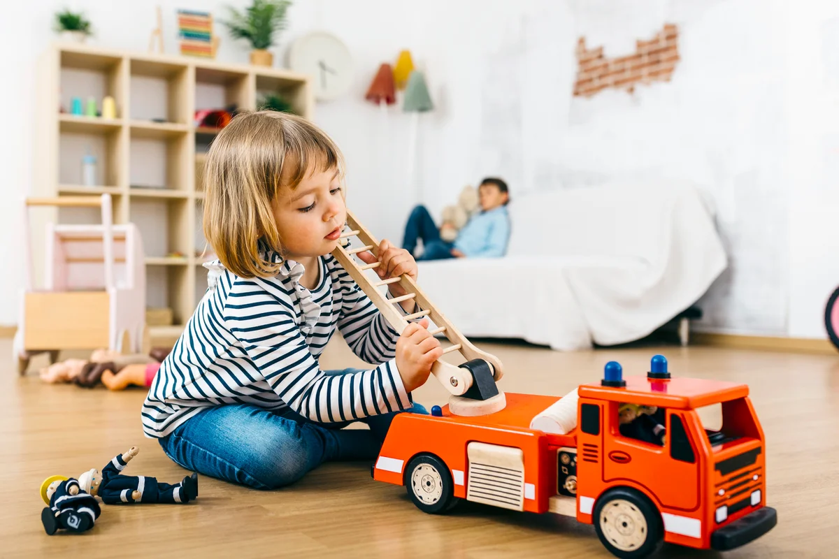 Girl playing with a wooden fire truck and brother waiting