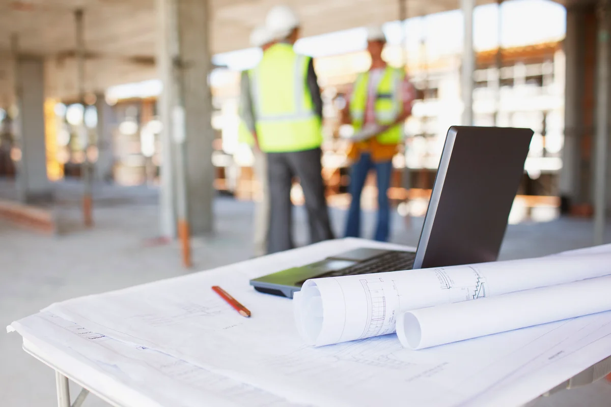 Construction workers behind blueprints and laptop on construction site