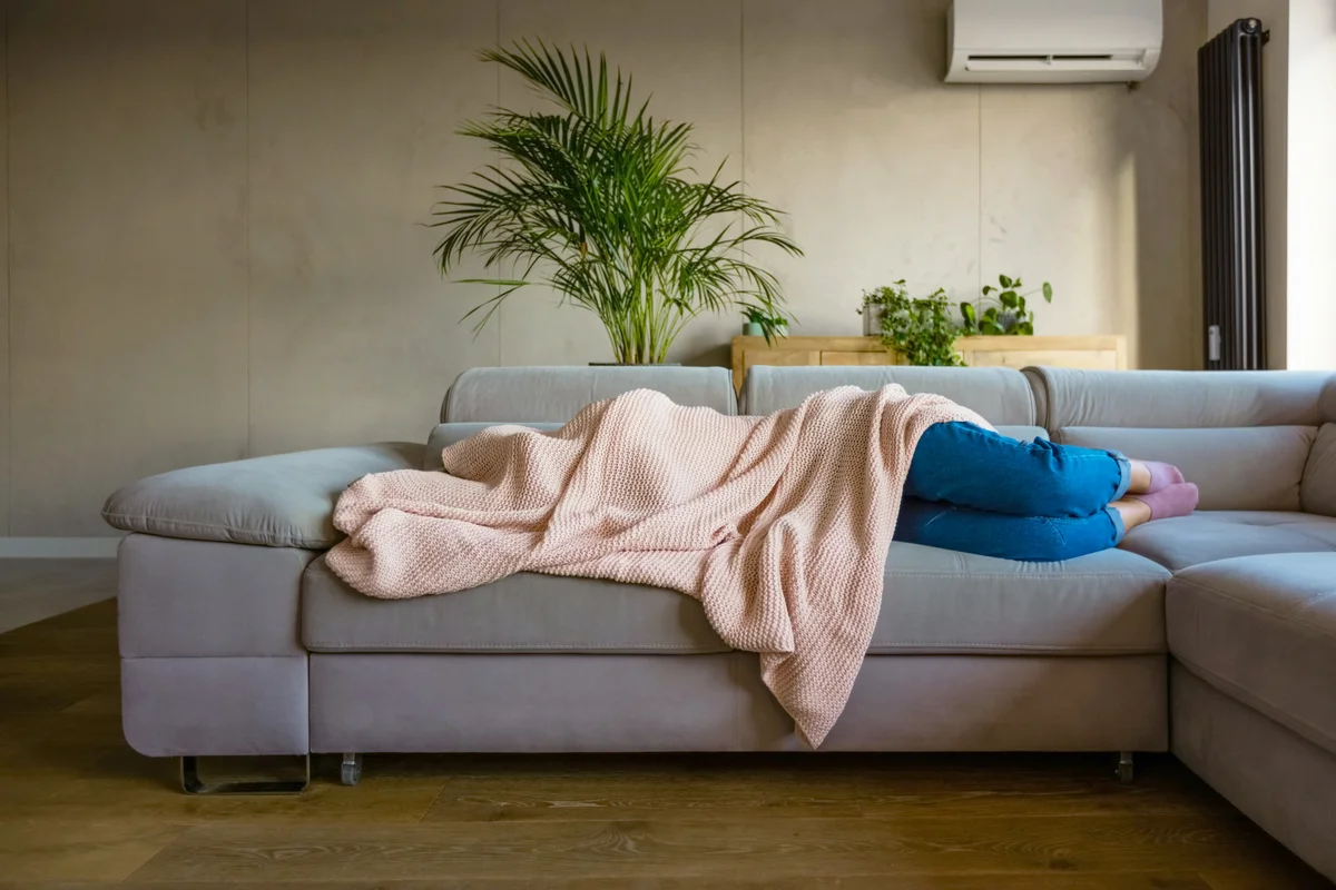 Young woman lying down on sofa in living room covered by blanket