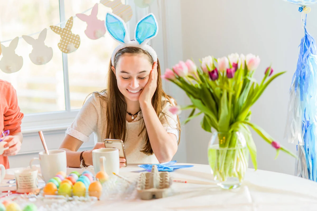 Teenage girl at table reading smartphone texts whilst dyeing hard boiled eggs for Easter