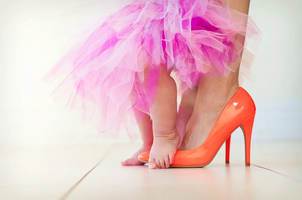 Baby with bare feet standing on high heels on white tile floor
