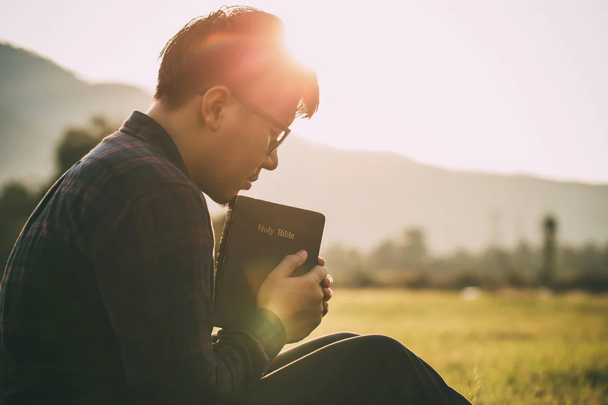 man praying on the holy bible in a field during beautiful sunset