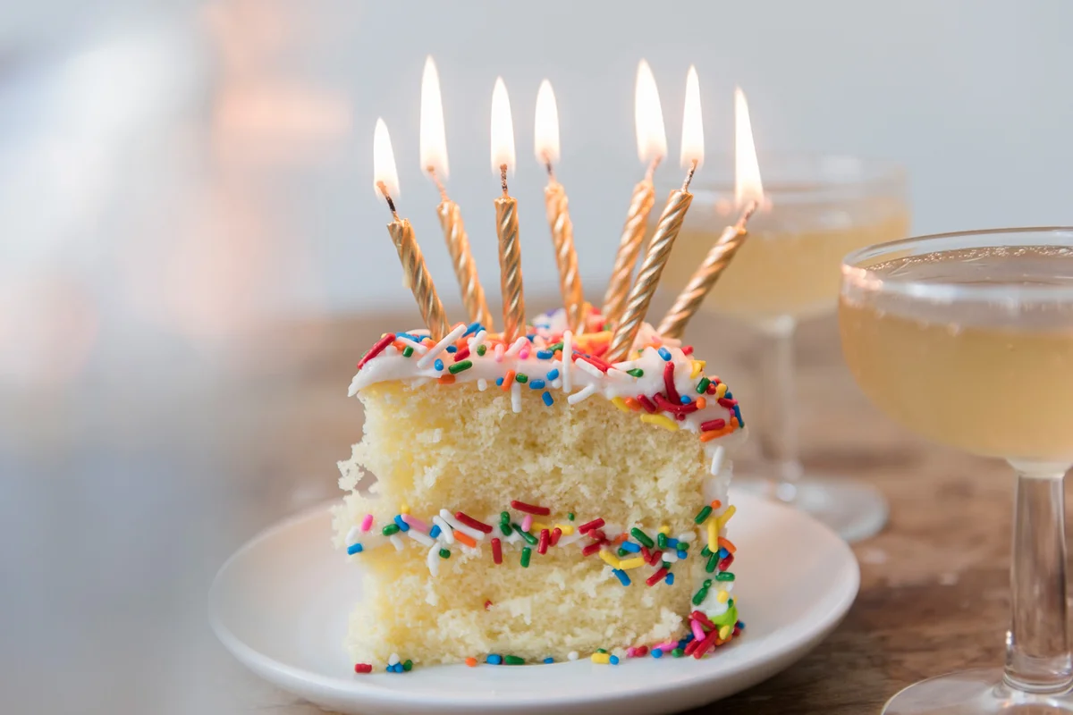 Candles burning on slice of cake with sprinkles near champagne